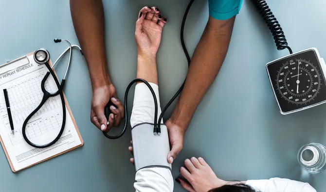 An arm is flat on a blue table with a doctor taking their blood pressure. There is a report file on a clip board next to them.