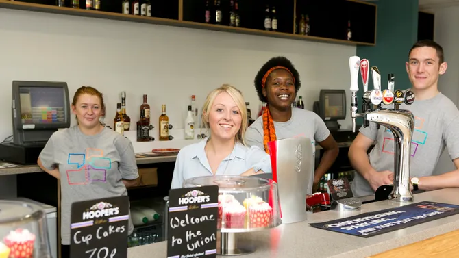 Staff working at the students' union are smiling from behind the bar.