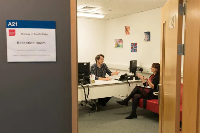 Two people sat, talking to eachother, in a therapy room. One is sat behind a desk and the other is sat on the sofa.