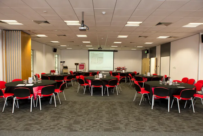 A collection of round tables containing glass bottles of water and cups surrounded by chairs in a smart conference room with a stage and a projector screen at the front