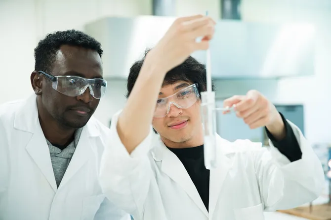 Two students in lab coats and protective eyewear are measuring liquid in a test tube.