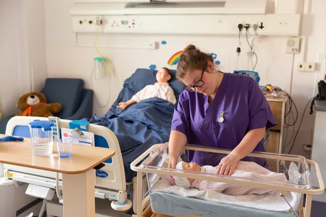 A student nurse wearing purple scrubs is caring for two mannequins that simulate a baby and a child in a simulated hospital environment.