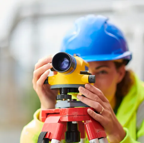 A person wearing a yellow high visibility jacket and a blue hard hat looks through a piece of surveying equipment