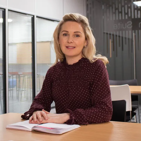 A HR student looks at camera while sitting at a wooden desk with their hands on top of a book in a meeting space