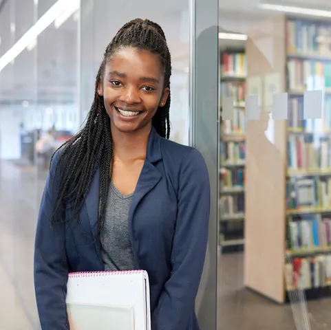 A student holding a notepad smiles at the camera while stood in the library in Treforest