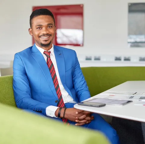 A student wearing a blue suit smiles at the camera while being sat on a green sofa at a desk with papers, a tablet and a phone in the USW Postgraduate Centre in Treforest