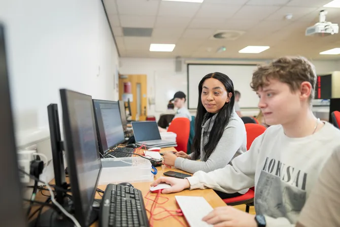 A student smiles while working with her course mate at a desktop computer in a computing  workshop at Treforest campus. There are other students working an academic helping in the background