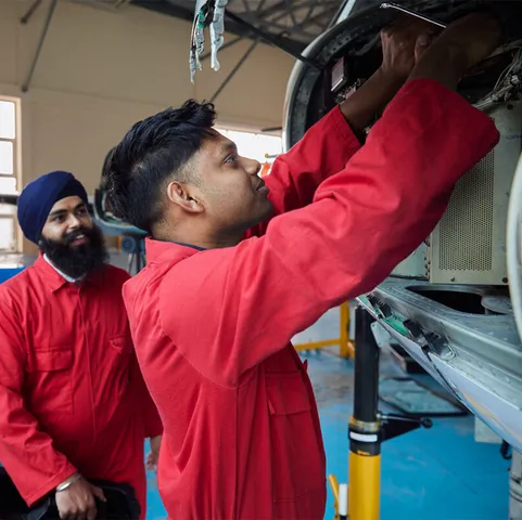 A student in red overalls repairs part of a small aircraft as another student watches