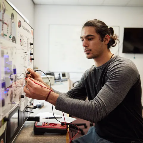 A postgraduate student conducts an experiment using wires and a electronic panel with small lights on a wall in a USW laboratory