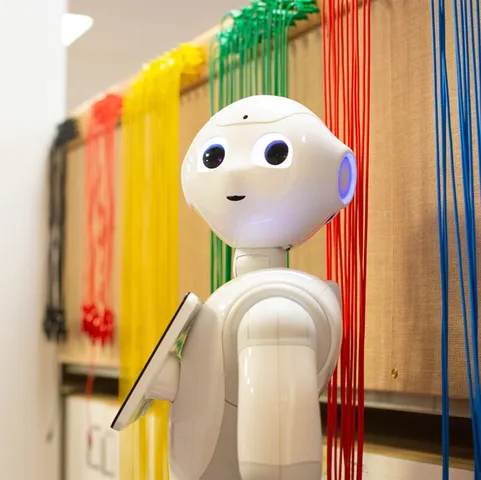 Pepper the robot looks at the camera in front of a wall covered in groups of wires separated into different colours in an en electrical engineering workshop at USW Treforest