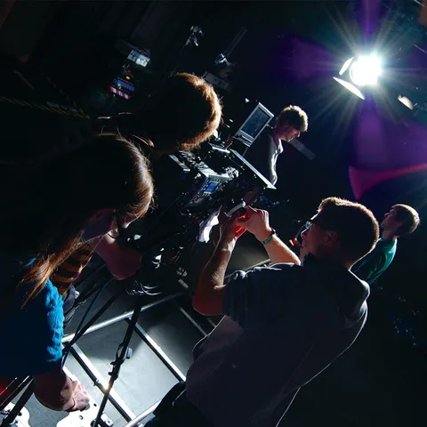 A group of students help to operate a camera filming two people chatting under a spotlight in a dark film studio