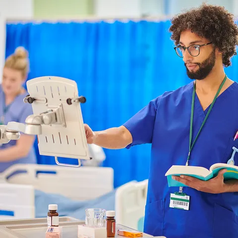 A nurse holding a textbook whilst inputting information on to a monitor