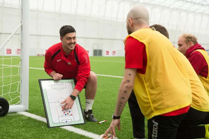 A student is communicating a game plan that is drawn on a small whiteboard to other students at the side of a football pitch.