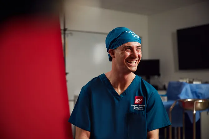 Student Noah Beuschel smiles and poses in front of a red backdrop and a USW simulation hospital ward while wearing medical scrubs and an ODP hair cap