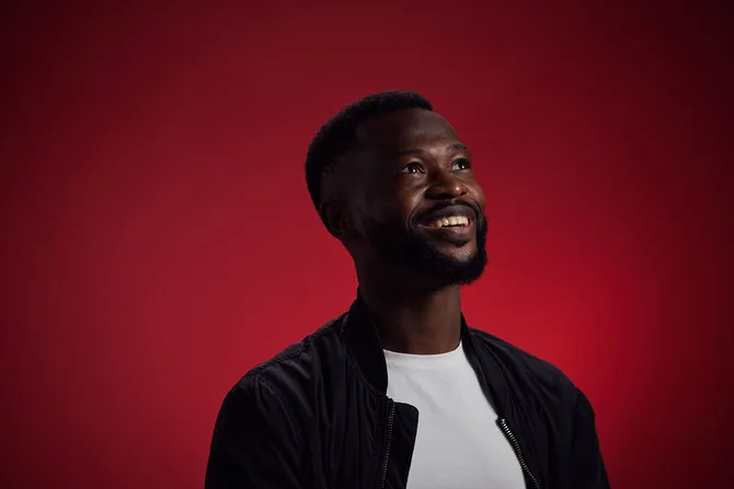 Alumni, Dezire Mambule, sat in front of red backdrop smiling looking at the ceiling