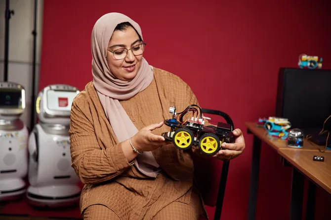 Senior Lecturer, Dr Mabrouka Abuhmida, holding a robotic car model in front of a red backdrop, with two small robots in the background
