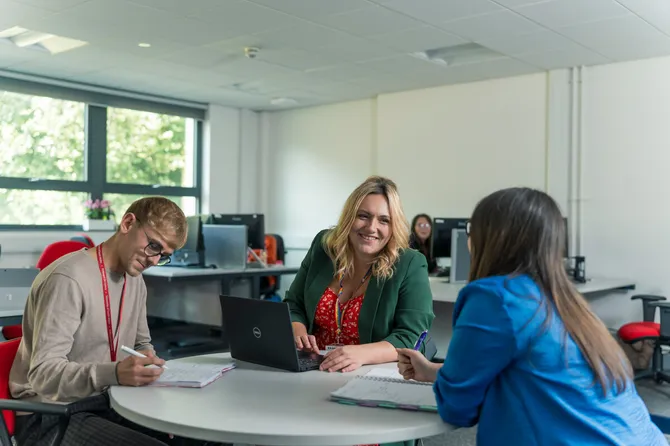 three members of the faculty of business and creative industries team sitting around a table having a meeting, taking notes in notebooks and a laptop
