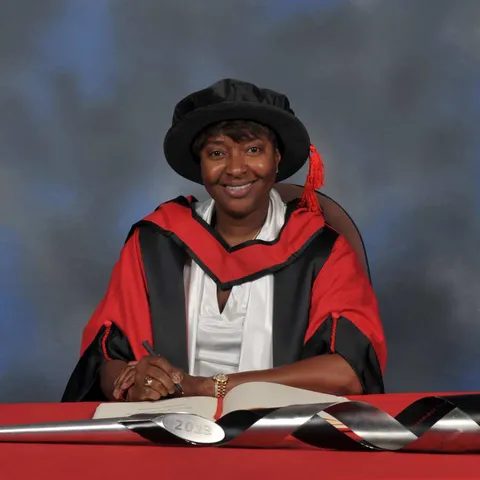 La Chun Lindsay writes in a large book next to a ceremonial mace on a red desk while wearing a doctorate's cap and gown in front of a blue backdrop.