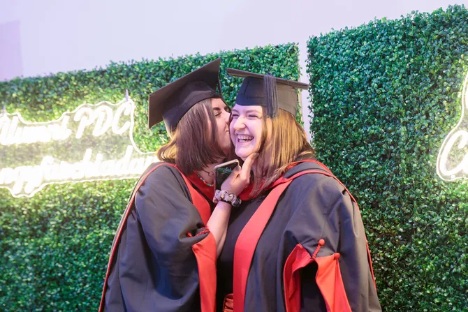 A student dressed in graduation robes and a graduation cap kisses another graduating student on the cheek in front of a leafy backdrop at Newport's ICC