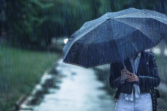 A person huddles under a black umbrella in heavy rain while walking on a tarmac path through a park
