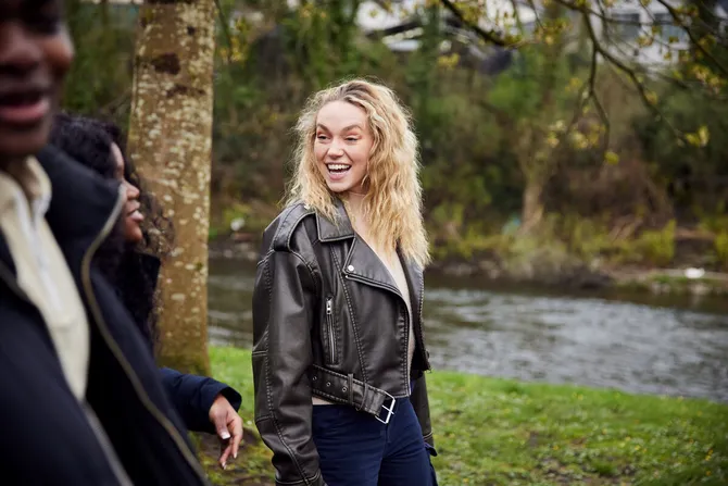 A student smiles while walking with their friends next to the grassy riverbank of the river Taff in Pontypridd