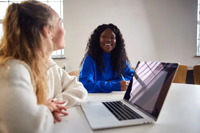 Two students studying and smiling. One is on a laptop and one is writing notes on paper.