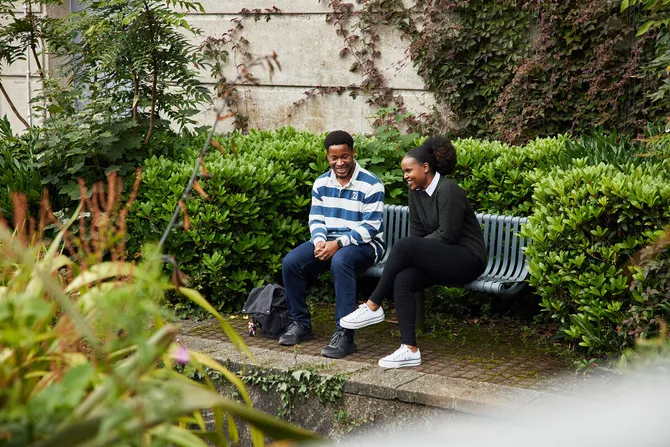 Two students sat outside on a bench surrounded by greenery.
