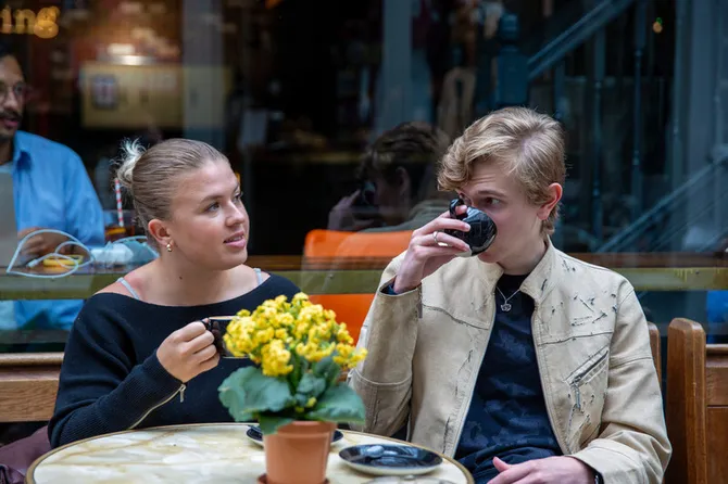 Two students sat outside a coffee shop. One is talking and the other is taking a sip of coffee.