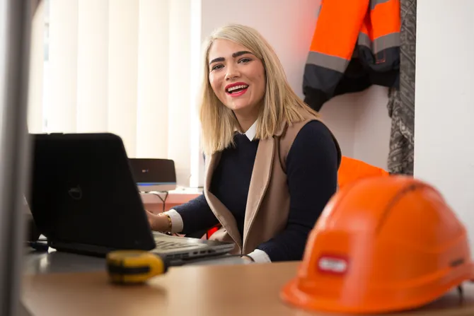Student, Mercedes, sat at a desk that has a computer, tape measure and a hard hat on it. She is working and smiling at the camera.