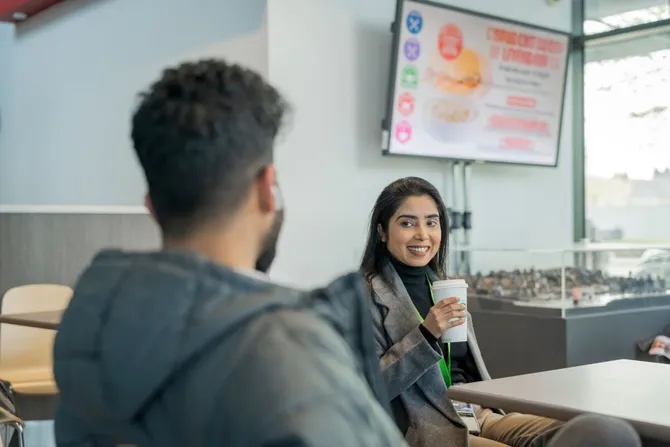 A cyber security student smiles at their friend and chats while drinking a coffee at Newport Campus
