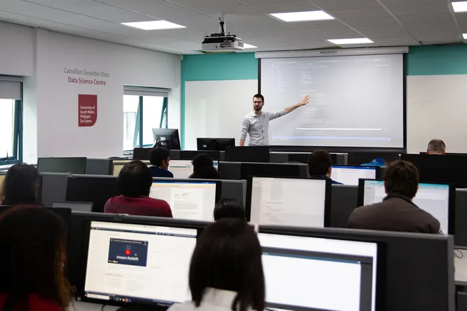 A data science classroom full of computers and students with a lecturer presenting at the front