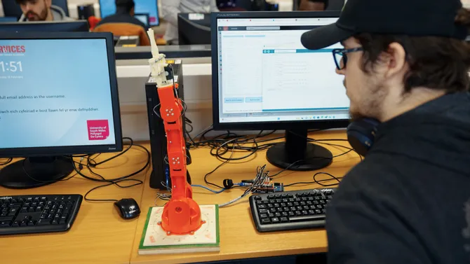 A student observes a robot arm with a claw on the end on a desk while sat at a computer at the Innovation Hub in an engineering workshop at the Treforest campus