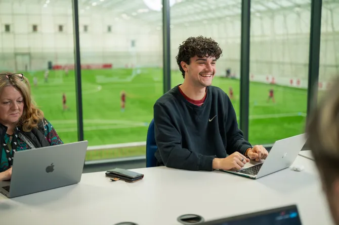 Sports Journalism student, Freddie, is sitting in a classroom with his laptop. There is a view of a football pitch through the glass windows behind him.