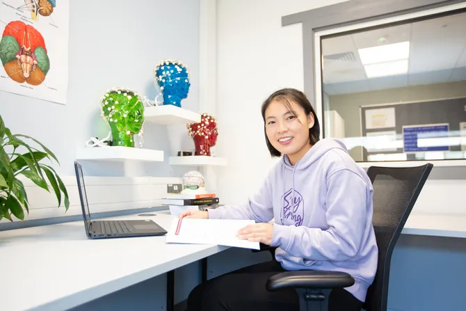 A student is sat at a desk in a Psychology lab, smiling into the camera.