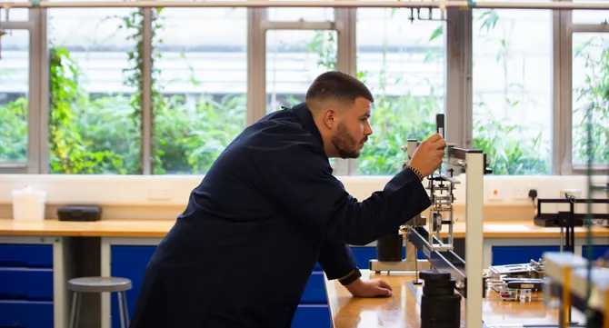 A student wearing navy engineering overalls adjusts the measurements on a piece of engineering equipment on top of a desk in a workshop at USW's Treforest campus. Behind the student is windows showing plants outside against a white wall