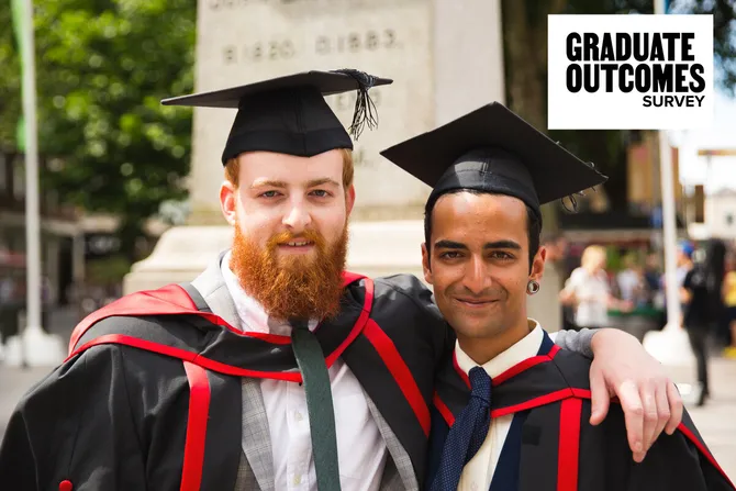 Two students wearing red and black graduation gowns and graduation caps smile with their arms around each other's shoulders