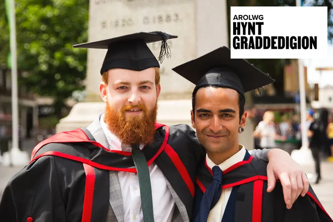 Two students wearing red and black graduation gowns and graduation caps smile with their arms around each other's shoulders