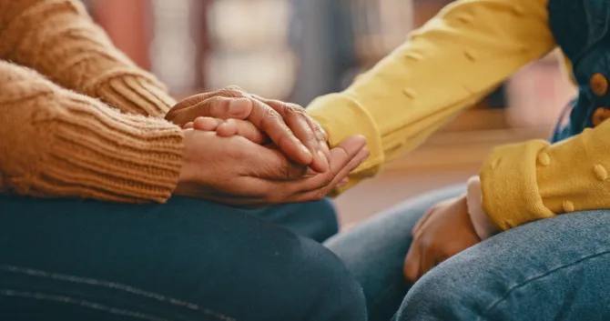 A close-up of two people holding hands while sat down in a comforting motion while talking
