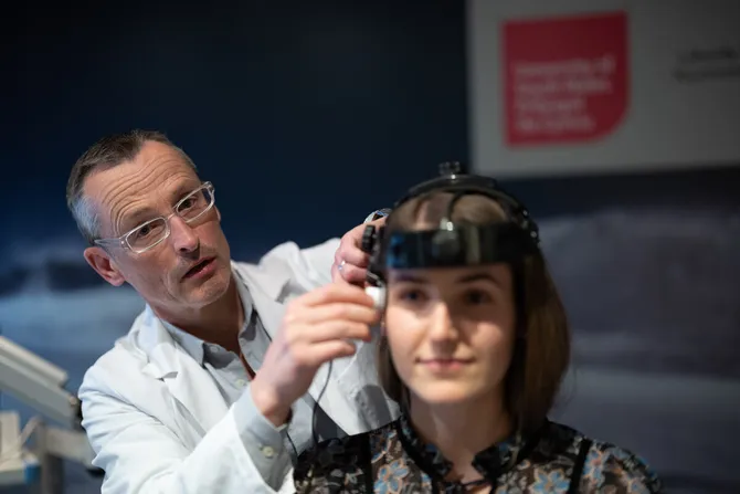 Professor Damian Bailey looks at the camera while dressed in a while laboratory coat as they adjust a piece of scientific equipment on a student's head
