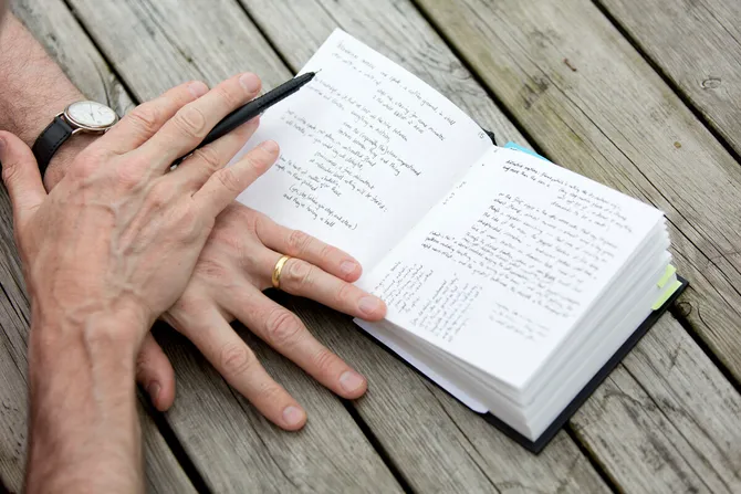 A pair of hands rest on top of each other while holding a pen and wearing a watch on top of a small notebook that is filled with handwritten notes on a picnic table
