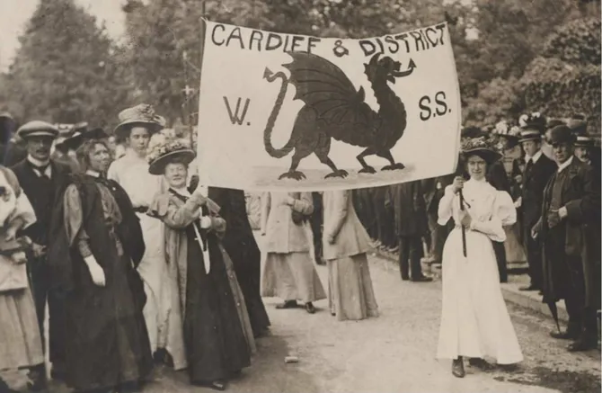 A historical black and white photo of women stood outside in front of trees holding a protest banner containing the Welsh Dragon, and the words 