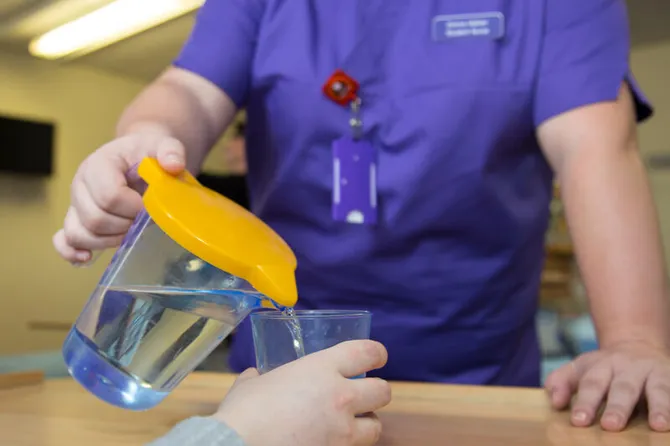 An adult nurse in purple scrubs pouring water into a plastic cup from a jug that has a yellow lid.