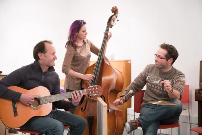 Three musicians smile while playing their instruments - including an acoustic guitar, a double bass and percussion instruments - together while sat down in front of a white wall