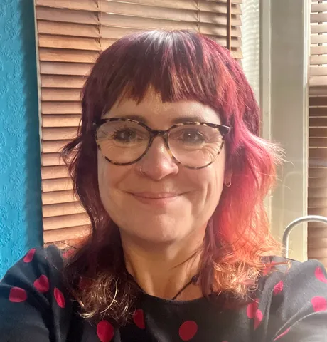 A woman with shoulder-length red and auburn hair, wearing glasses with a black frame and a black top with red polka dots, smiles at the camera. She has a nose ring and is sitting in a well-lit room with wooden blinds and a blue wall in the background.