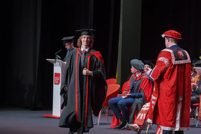 An undergraduate student walks across a stage in front of robed academics while wearing robes and an academics cap to graduate from their degree at the ICC in Newport