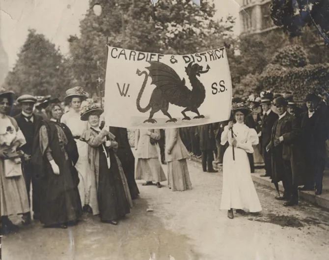 Black and white photo of early 20th-century suffragists marching in a demonstration. A group of women and a few men are gathered on a street, with two women holding a large banner that reads “Cardiff & District W.S.S.” featuring a prominent Welsh dragon.