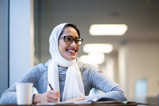 A woman wearing a hijab is sat writing notes at a desk and smiling.