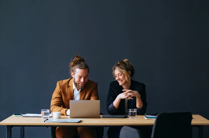 Smiling businessman and businesswoman using a laptop computer while talking and sitting together at office desk.