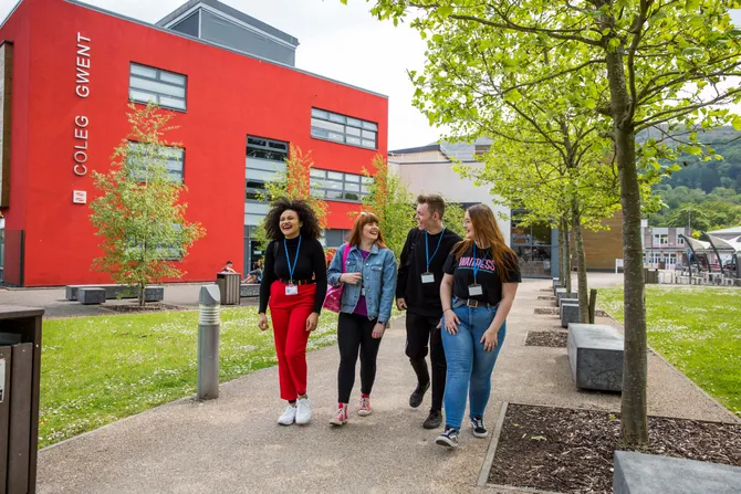 Four students walking past a red college painting with the Coleg Gwent logo on it.