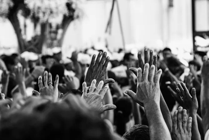 A black and white photo of a crowd of people raising their hands in unison.